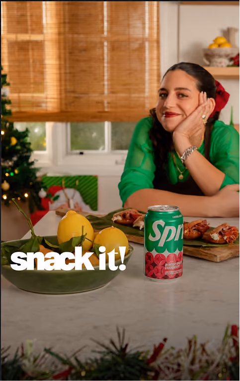 A woman with dark hair tied back with a red ribbon, wearing a green top, leans on a kitchen counter smiling. The counter has a green bowl of lemons, a can of Sprite cranberry flavor, and a wooden tray with appetizers. Behind her is a window with bamboo blinds and a decorated Christmas tree.
