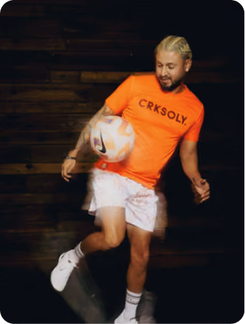 Man with blond hair wearing an orange CRKSOLY shirt, white shorts, and white sneakers, juggling a soccer ball against a dark wooden background.