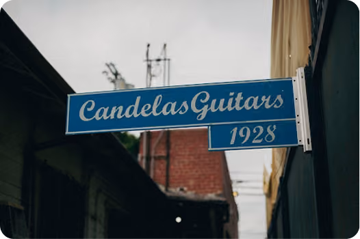 Blue rectangular sign with white cursive text reading 'Candelas Guitars' and the year '1928' below it, mounted on a wall outside between buildings under a cloudy sky.