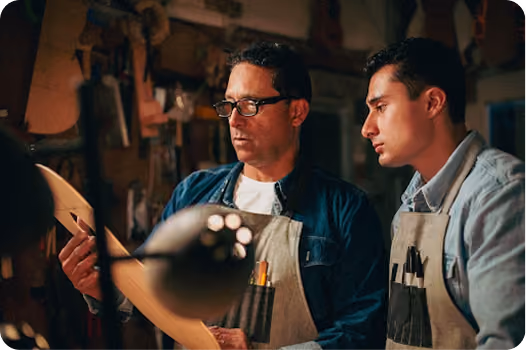 Two men wearing aprons in a workshop are examining a piece of wood or paper, focused on their craft.