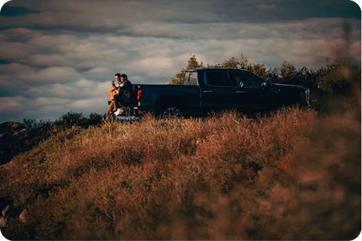 Two people sitting on the lowered tailgate of a dark pickup truck parked on a grassy hill under a cloudy sky.
