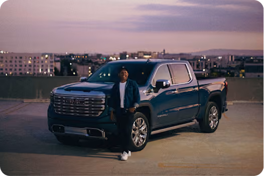 Man wearing a dark jacket and white sneakers leaning against the front side of a dark blue pickup truck parked on a rooftop with a cityscape in the background at dusk.