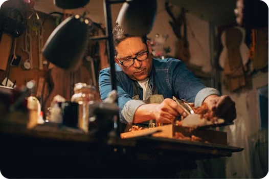 Man wearing glasses and a denim jacket working intently with a wood plane on a piece of wood in a dimly lit workshop filled with tools and guitars.
