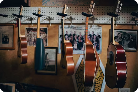 Six acoustic guitars hanging by their necks on a pegboard wall with framed photos in the background.