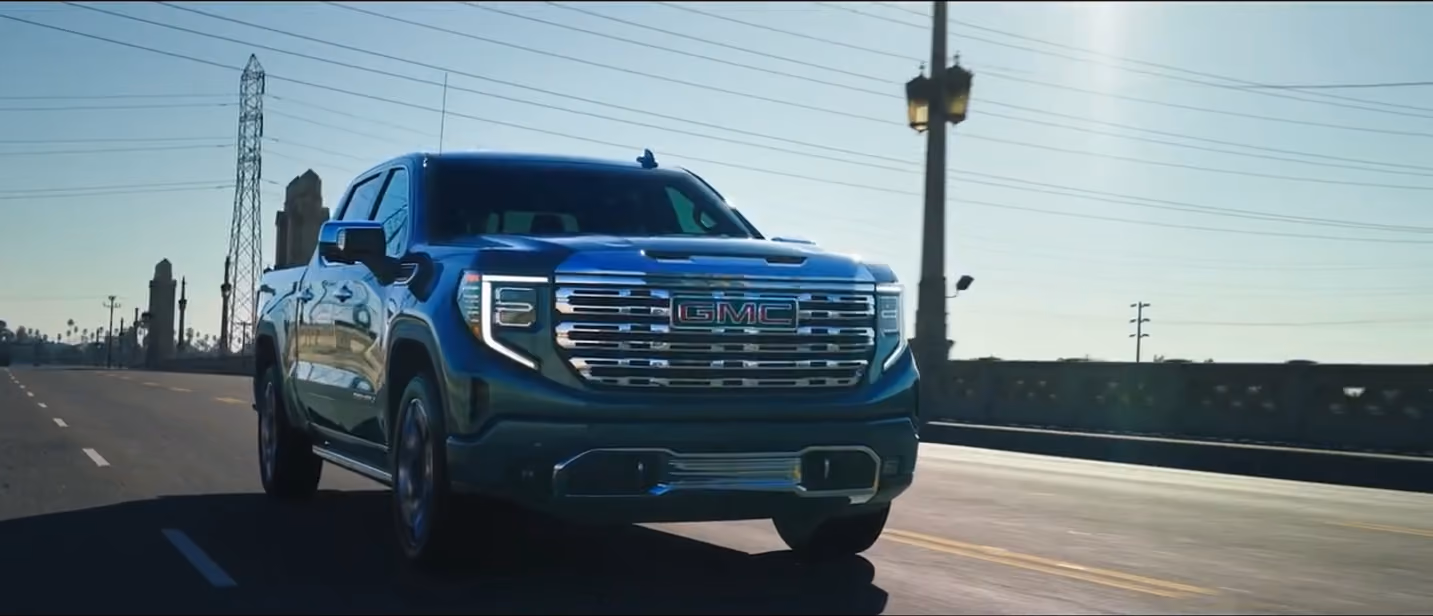 Dark green GMC pickup truck driving on an empty road with power lines and towers in the background during daylight.