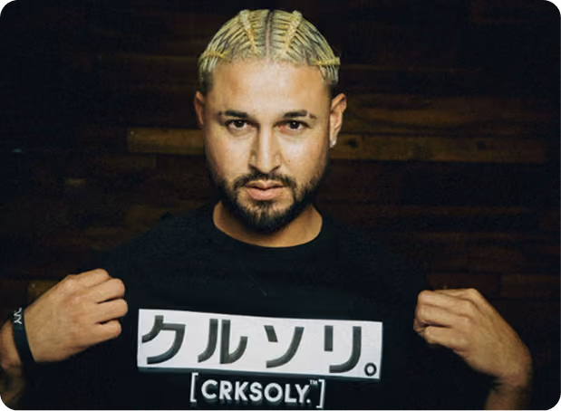 Man with braided blonde hair and beard wearing a black shirt with white Japanese text and CRKSOLY branding, holding the shirt at the chest.