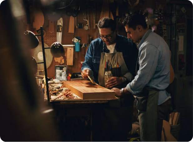 Two men wearing aprons working together on a woodworking project in a dimly lit workshop, applying finish or glue to a wooden board on a workbench cluttered with tools and wood shavings.