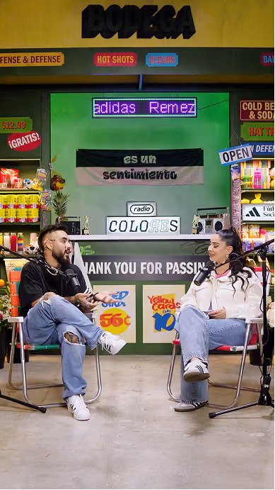 Two people sitting on chairs facing each other with microphones, recording a podcast or interview in a colorful bodega-themed studio with shelves of products, neon signs, and a banner reading 'es un sentimiento' and 'radio colores.'