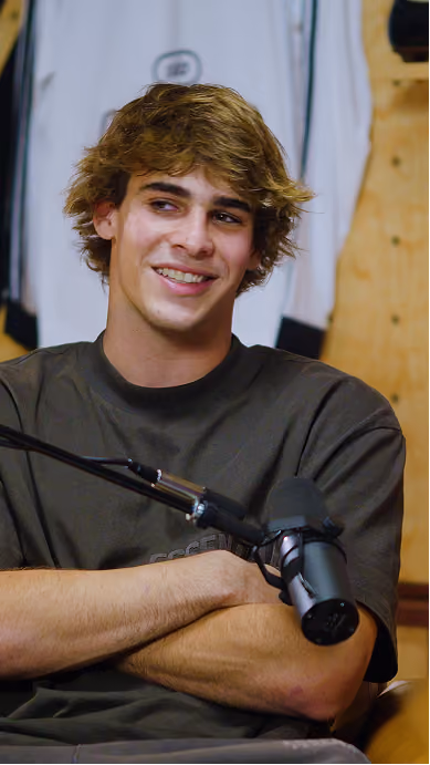 Young man with tousled brown hair smiling and sitting with crossed arms in front of a microphone in a casual indoor setting.