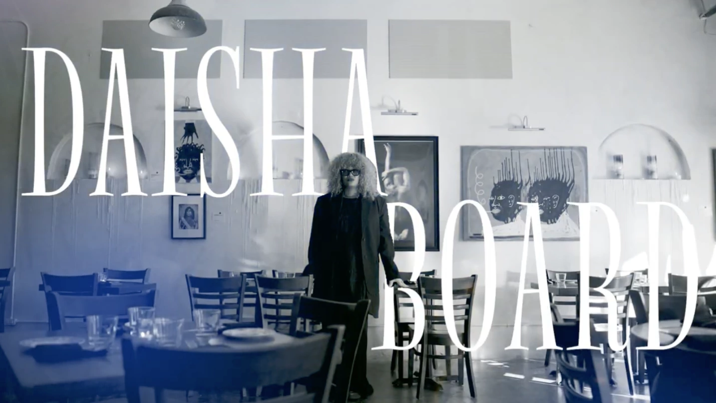 Person with curly hair and sunglasses standing in a restaurant dining area with wooden chairs and tables, with large text overlay reading 'DAISHA BOARD'.