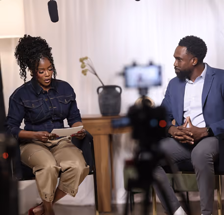 Two people seated facing each other in a studio setting with recording equipment visible; a woman on the left is reading from a paper and a man on the right listens attentively, a soft background with a wooden table and decorative plant behind them.