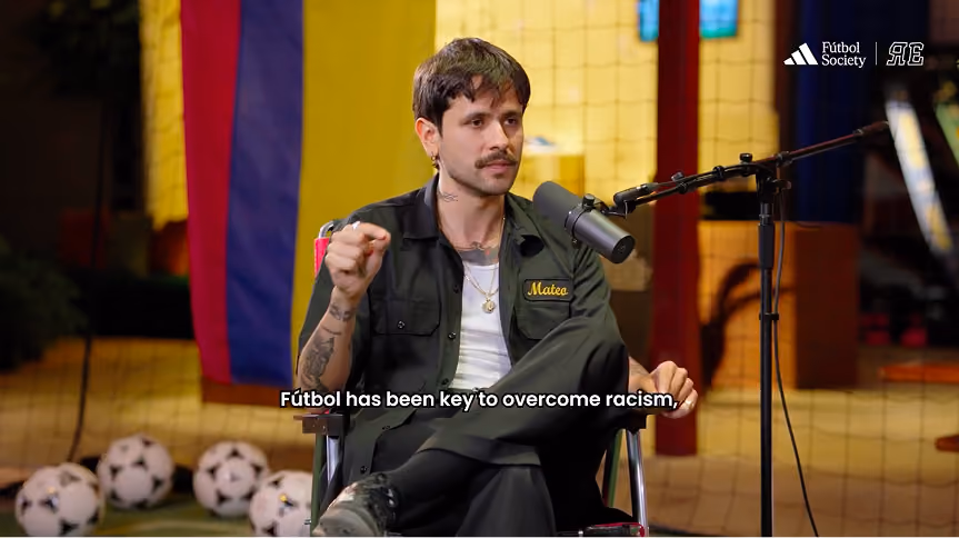 Man with mustache and tattoos sitting on a chair in front of a microphone, speaking in a studio with a net backdrop and multiple soccer balls on the floor.