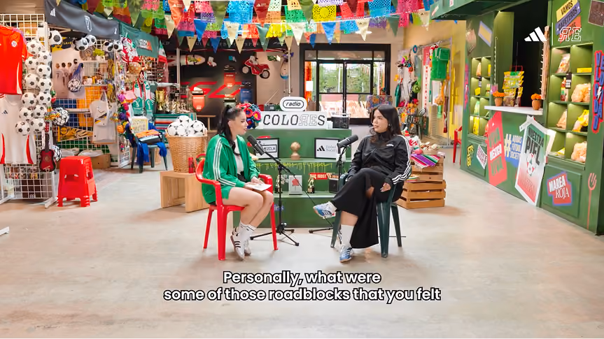 Two women seated facing each other in a colorful room decorated with soccer-themed items, pennant flags hanging from the ceiling, and shelves displaying various merchandise; one woman wears a green jacket and white sneakers, the other a black jacket and blue sneakers, speaking into microphones.