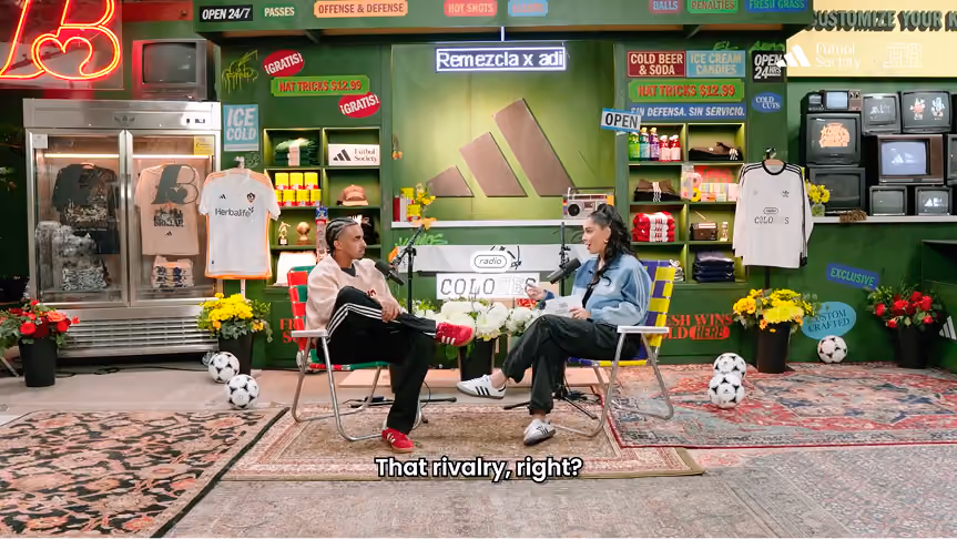Two people sit facing each other on metal chairs in a colorful room decorated with soccer memorabilia including jerseys, soccer balls, flowers, vintage TVs, and neon signs; one person is wearing a light jacket and red shoes, the other is in a blue jacket and white shoes, with a subtitle reading 'That rivalry, right?'.