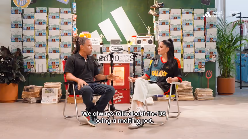 Two people seated and having a conversation in a studio with microphones; one person wears a black polo shirt and jeans, and the other wears a black Galaxy sports jersey and white pants. The background has colorful cards on green walls and a large white Adidas logo.