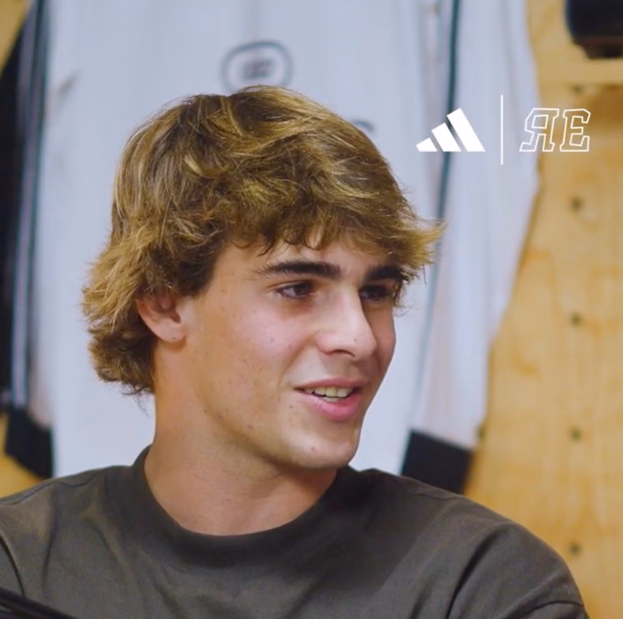 Young man with light brown hair smiling, wearing a dark shirt, with blurred sports jerseys hanging in the background and an Adidas logo visible.