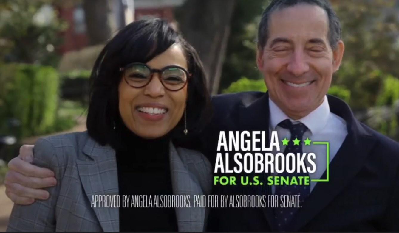 Angela Alsobrooks smiling and wearing glasses and a plaid blazer stands next to a smiling man in a suit and tie outdoors, with text beside them that reads "Angela Alsobrooks for U.S. Senate" and a disclaimer stating approval and payment by Alsobrooks for Senate.
