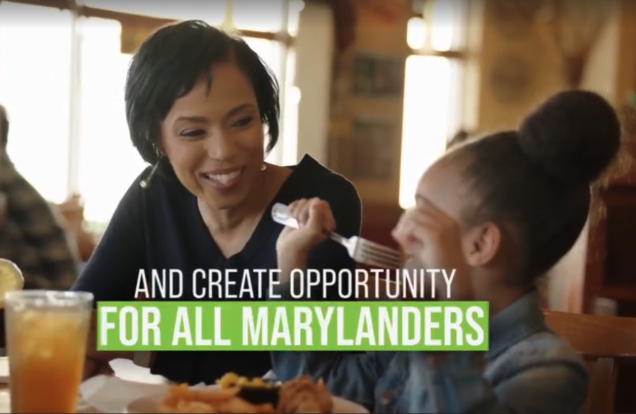 A smiling woman looks at a young girl with a fork in her hand as they sit together at a table with food and a glass of iced tea in a warmly lit restaurant, with overlaid text saying 'AND CREATE OPPORTUNITY FOR ALL MARYLANDERS'.
