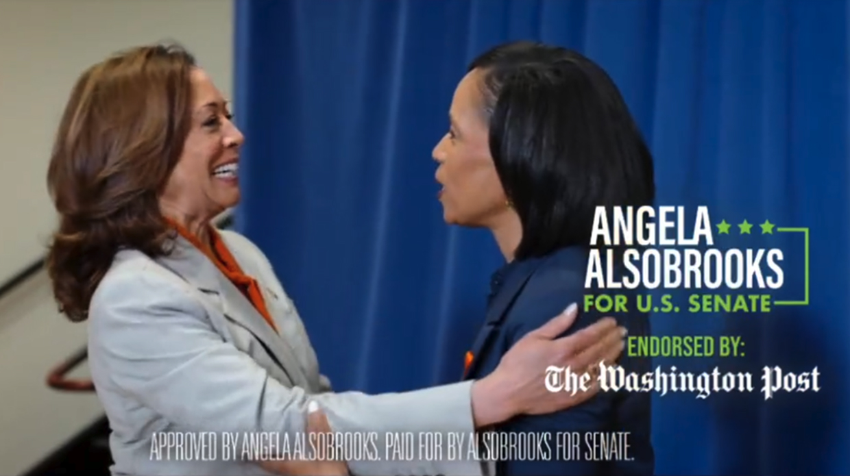 Two women warmly greeting each other by holding arms and smiling, with a blue curtain background and campaign text that reads: Angela Alsobrooks for U.S. Senate endorsed by The Washington Post.