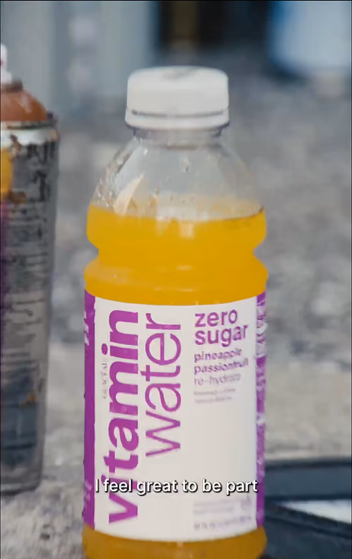 A clear plastic bottle of vitaminwater with orange liquid inside, labeled 'zero sugar pineapple passionfruit' on a white and purple label, on a textured gray surface with blurred background.