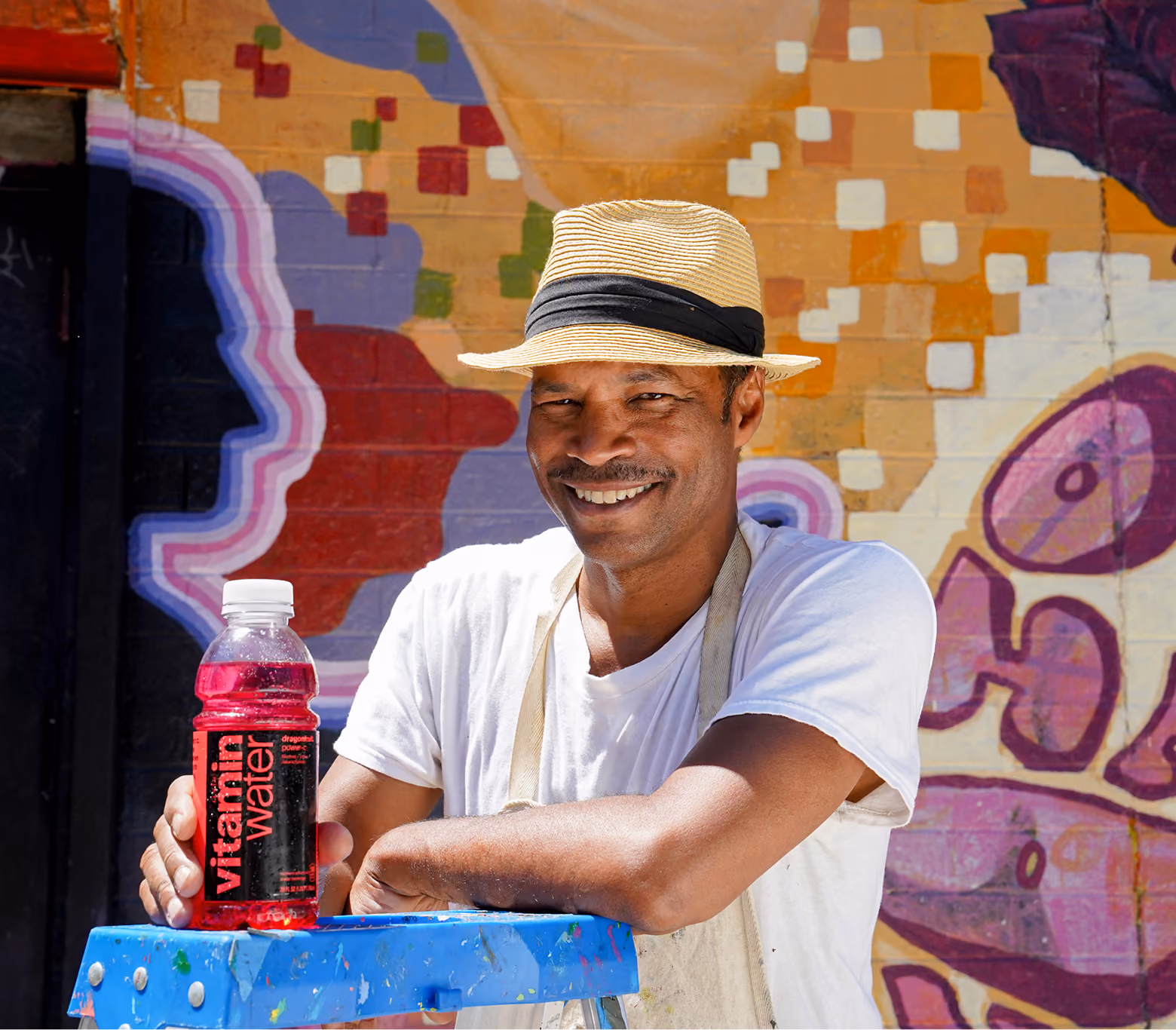 Smiling man wearing a straw hat and white shirt leaning on a blue paint-splattered step stool, holding a red bottle of vitaminwater, with a colorful graffiti wall in the background.
