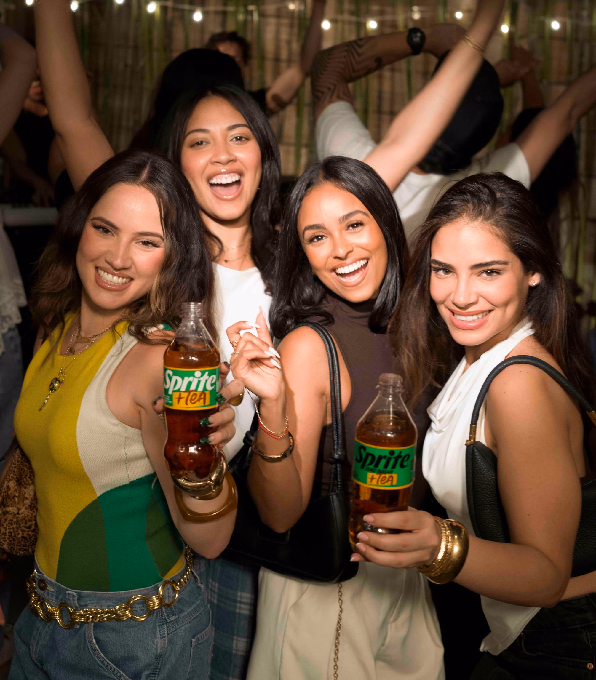 Four young women smiling and posing at a party, two holding bottles of Sprite + Tea, with festive string lights in the background.