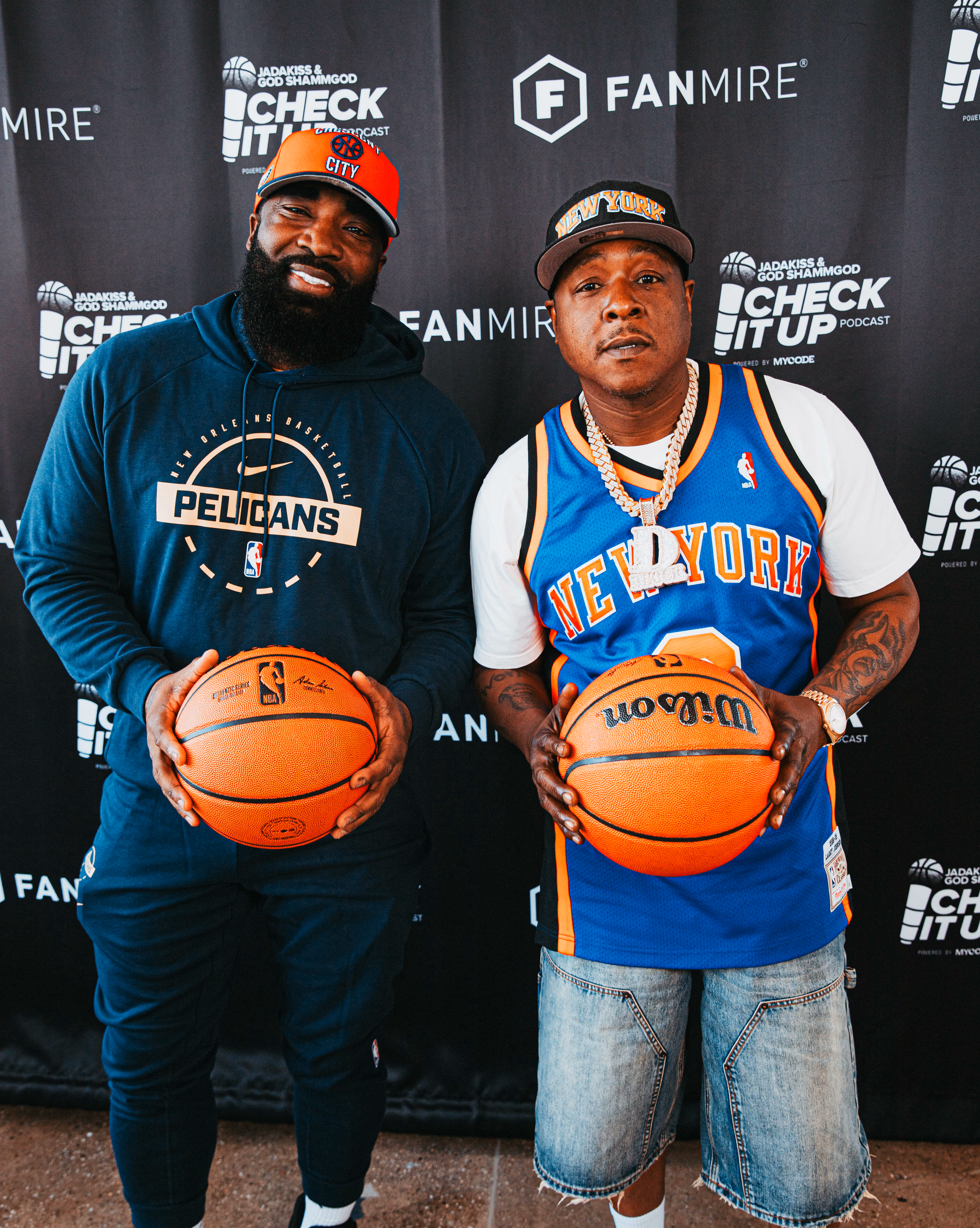 Two men holding basketballs stand in front of a backdrop with logos for the Check It Up podcast and Fanmire; the man on the left wears a New Orleans Pelicans hoodie and an orange hat, and the man on the right wears a blue New York Knicks jersey, denim shorts, a New York cap, and a large chain necklace with a 'D' pendant.