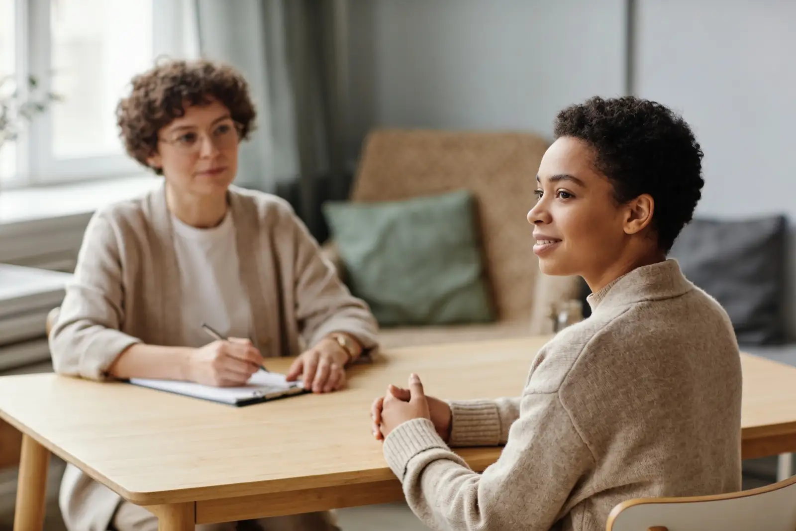 Two women sit at a wooden table in a counseling session, one listening attentively while the other speaks and smiles.