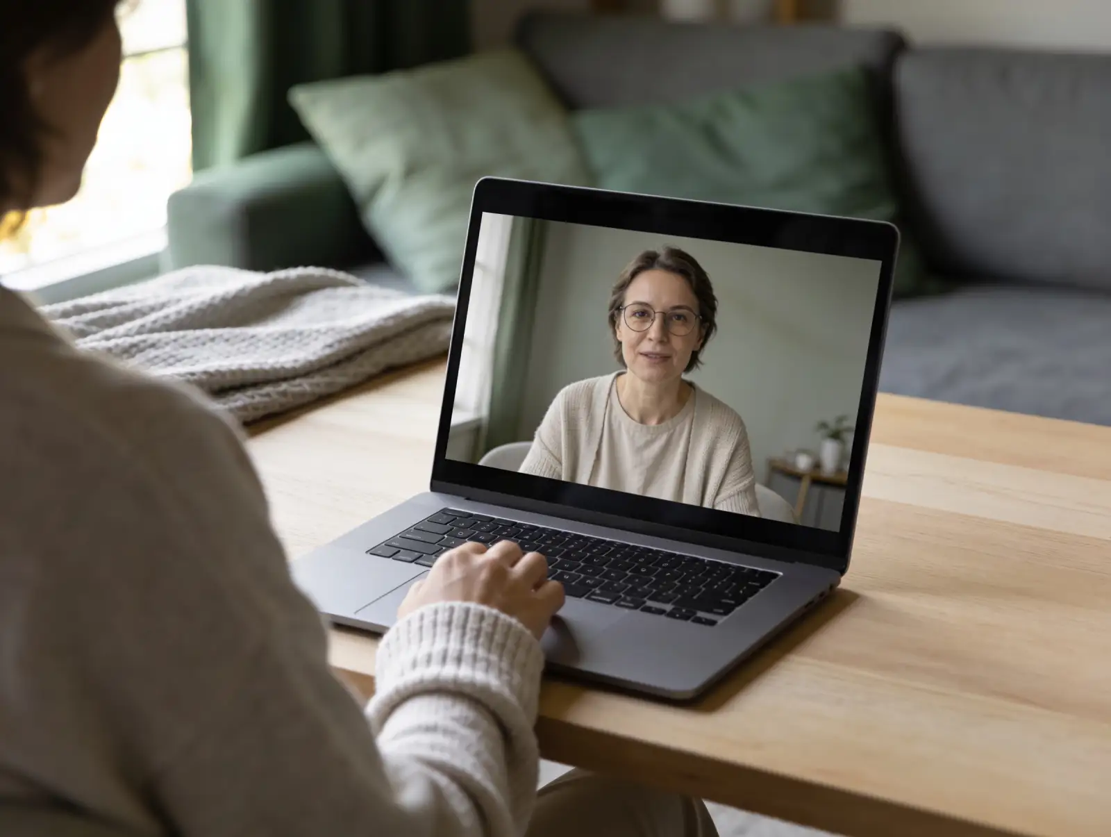Person with curly hair in a light sweater sitting at a wooden table, engaging in a video call on a laptop showing a woman with short brown hair, glasses, and a beige cardigan, in a softly lit room with neutral decor.