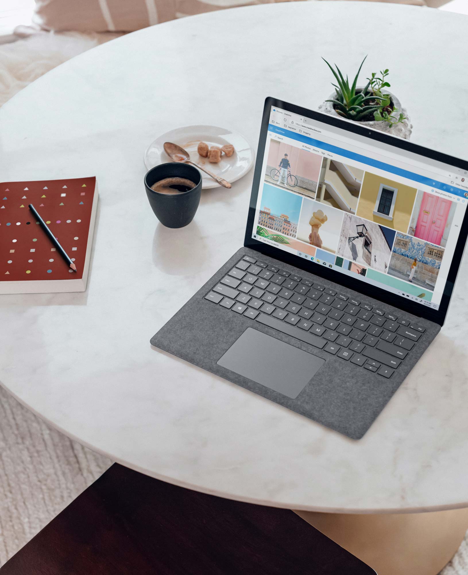 Open laptop on a round white marble table displaying a photo gallery on the screen, with a small potted plant, a black coffee cup, a plate with sugar cubes and a spoon, and a red notebook with a pencil beside it.
