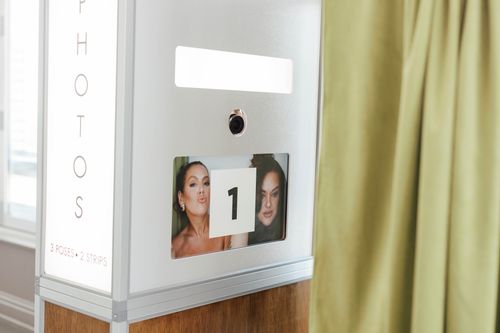 Photo booth machine with a screen displaying two women posing for a photo and the number 1 on a card in front of them, next to a green curtain.