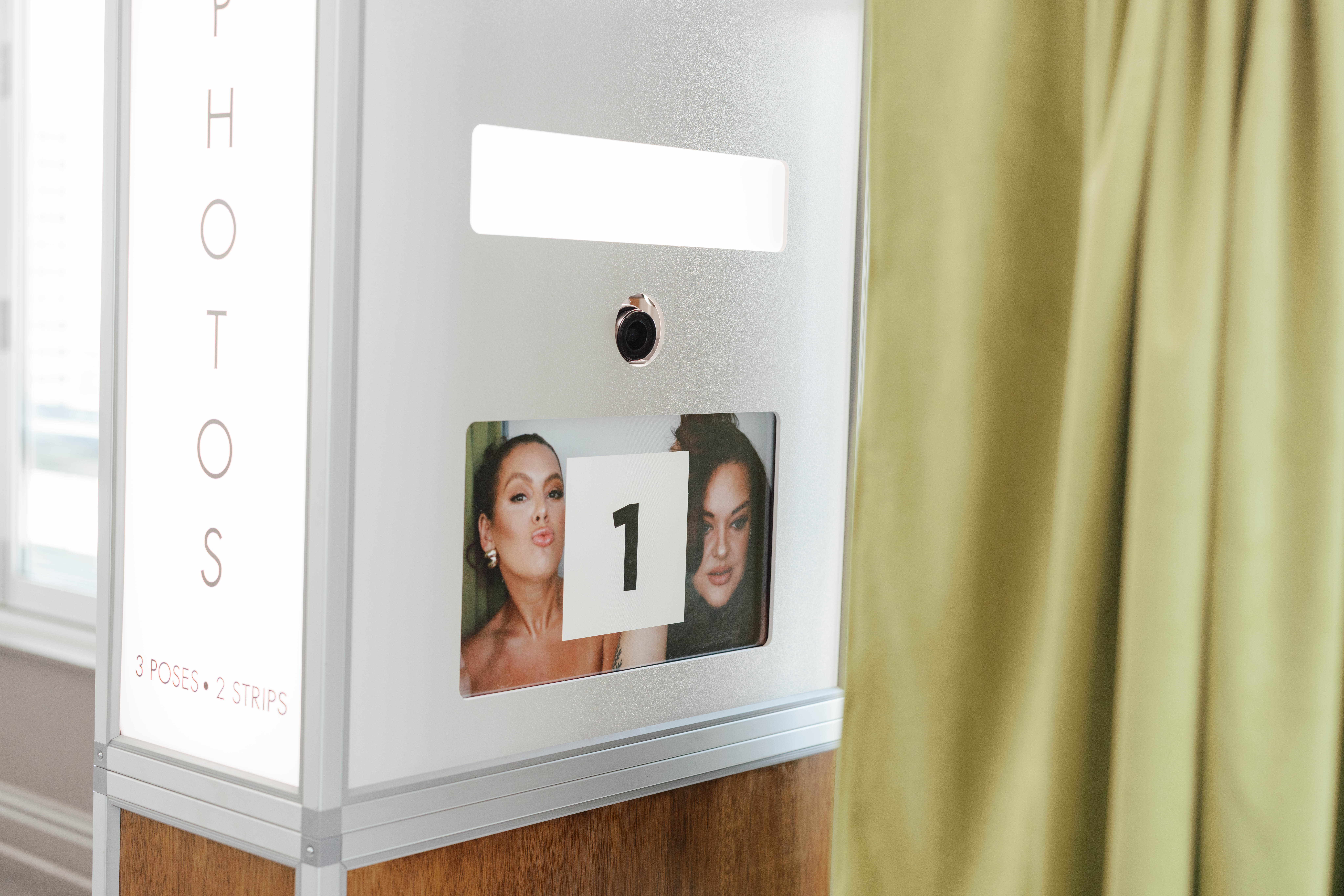 Photo booth machine with a screen displaying two women posing for a photo and the number 1 on a card in front of them, next to a green curtain.
