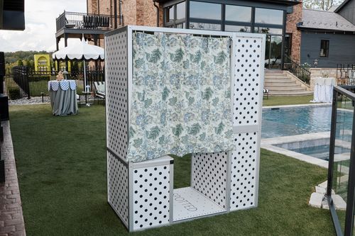 White rectangular photo booth structure with black polka dots and floral curtain in the entrance, placed on green grass near a swimming pool and a brick house.
