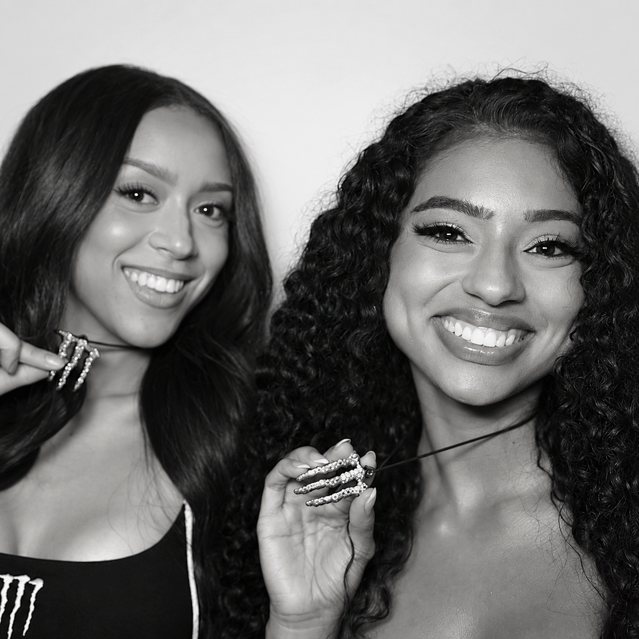 Two smiling women with long hair posing closely together, each holding a necklace pendant shaped like a claw with rhinestones. It is the logo pendant necklace for the energy drink brand, Monster. 
