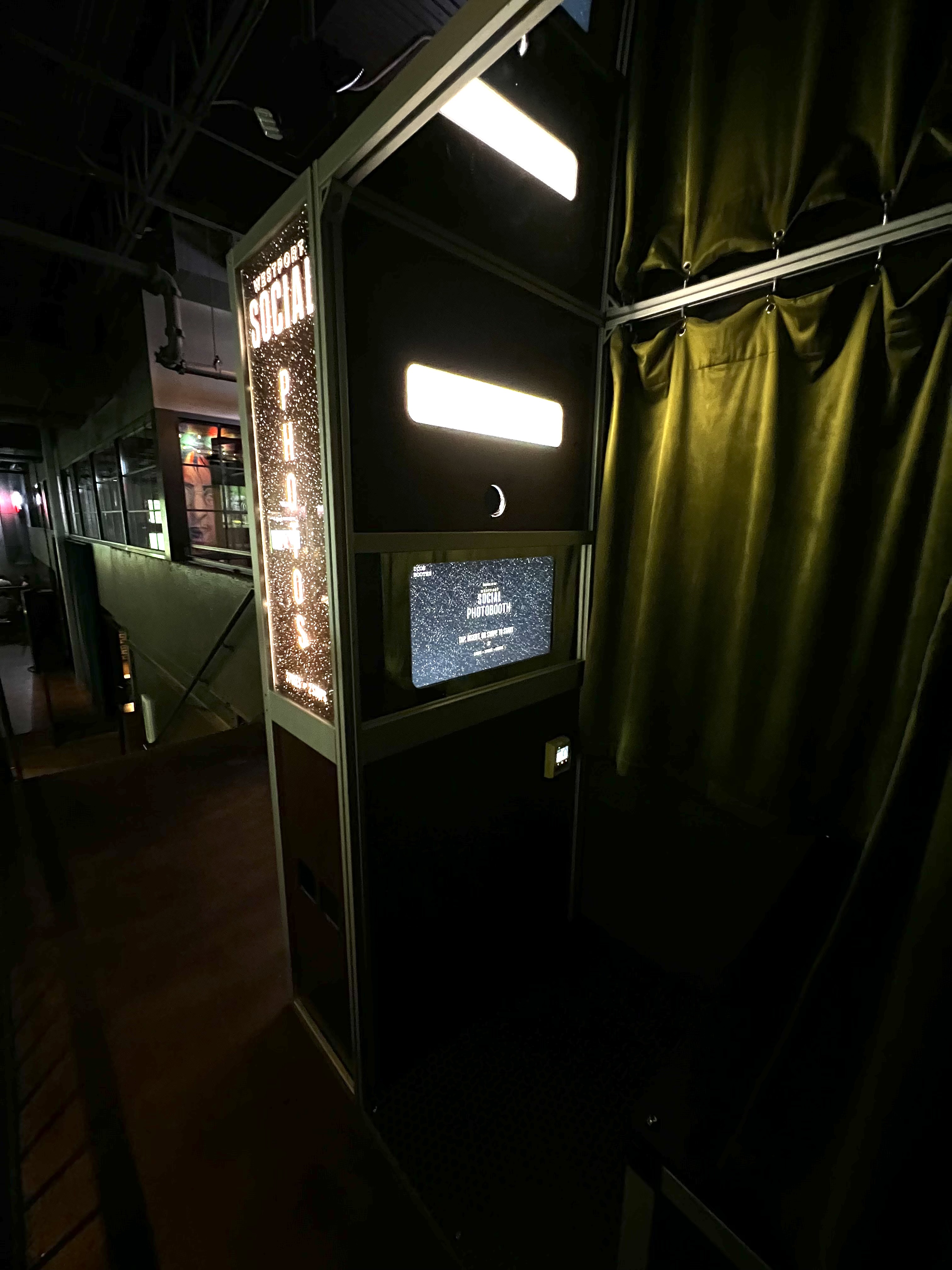 Dimly lit photo booth with illuminated sign reading 'Westport Social Photobooth' and a screen displaying instructions, next to green curtains.