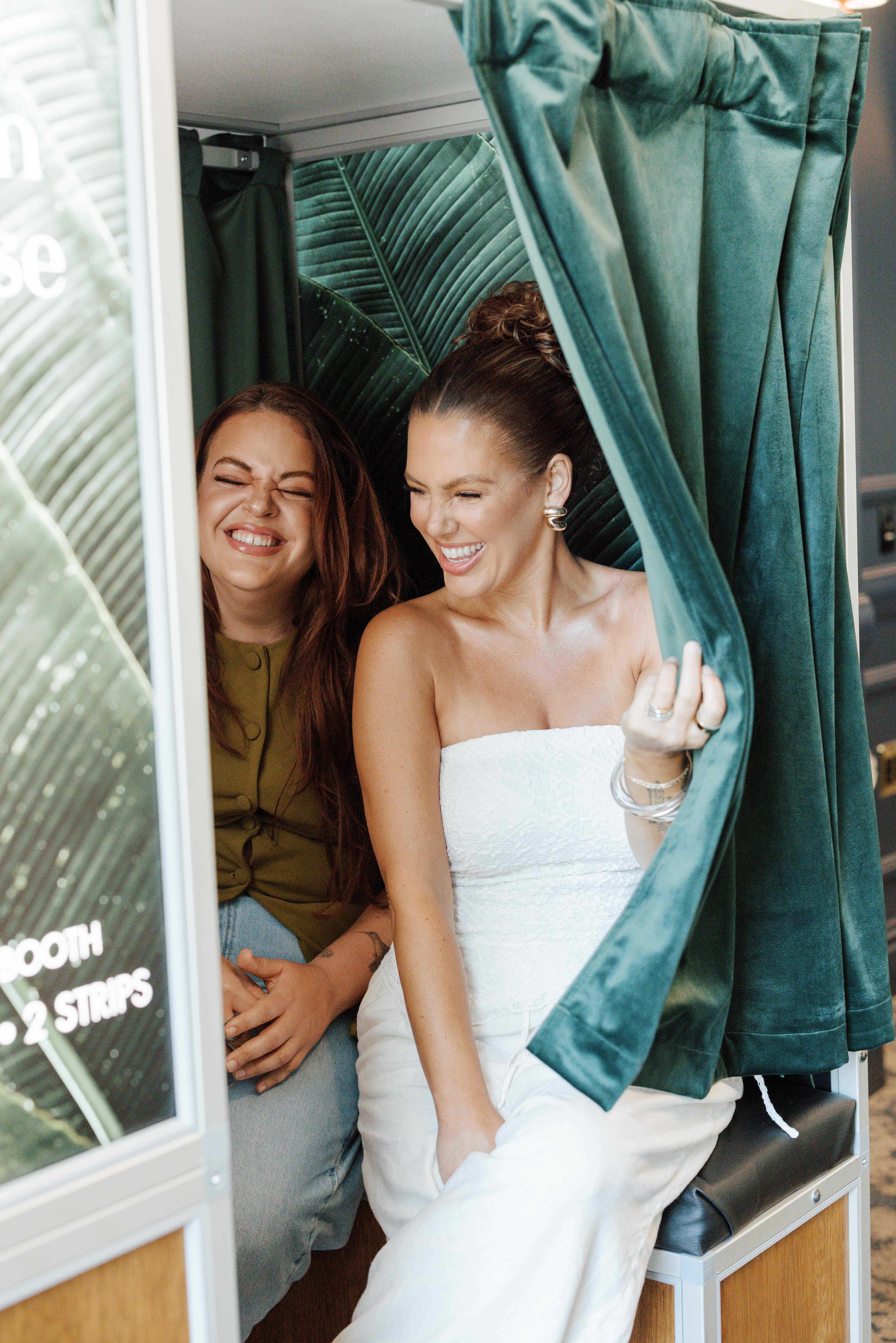 Two women sitting closely and laughing inside a photo booth with green velvet curtains and a tropical leaf backdrop, one woman pulling the curtain aside.