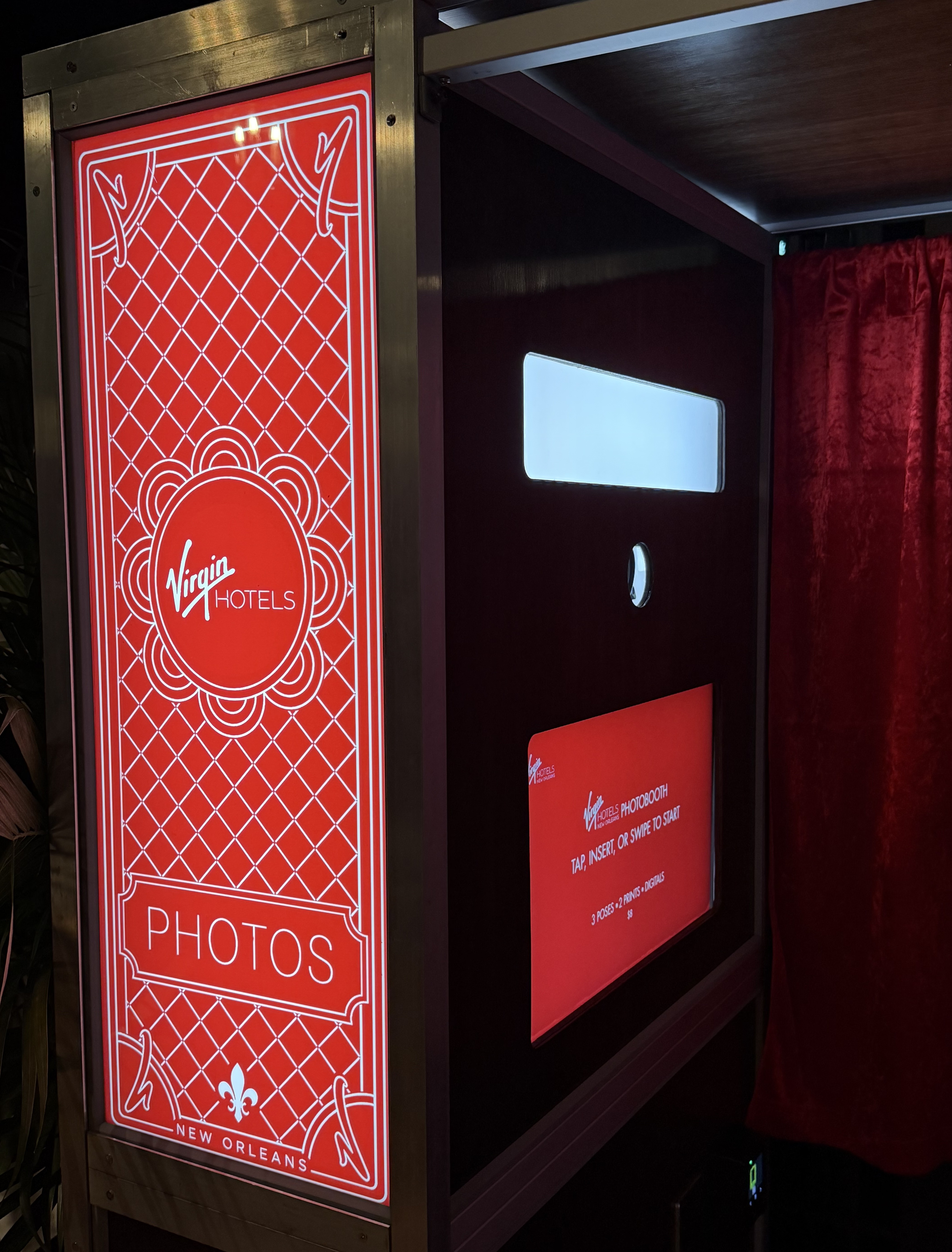 Virgin Hotels New Orleans photo booth with illuminated red panels, a touchscreen display, camera lens, and red velvet curtain.