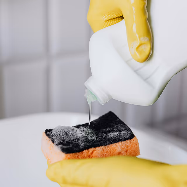 Person wearing yellow gloves pouring liquid detergent onto a black and orange cleaning sponge.