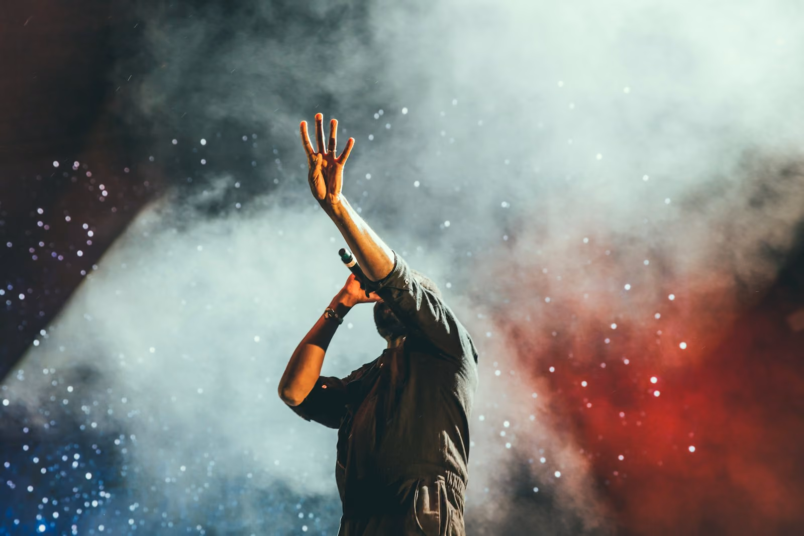 A male singer on stage holding a microphone in one hand with the other hand raised, surrounded by dramatic fog and colorful lights in the background.