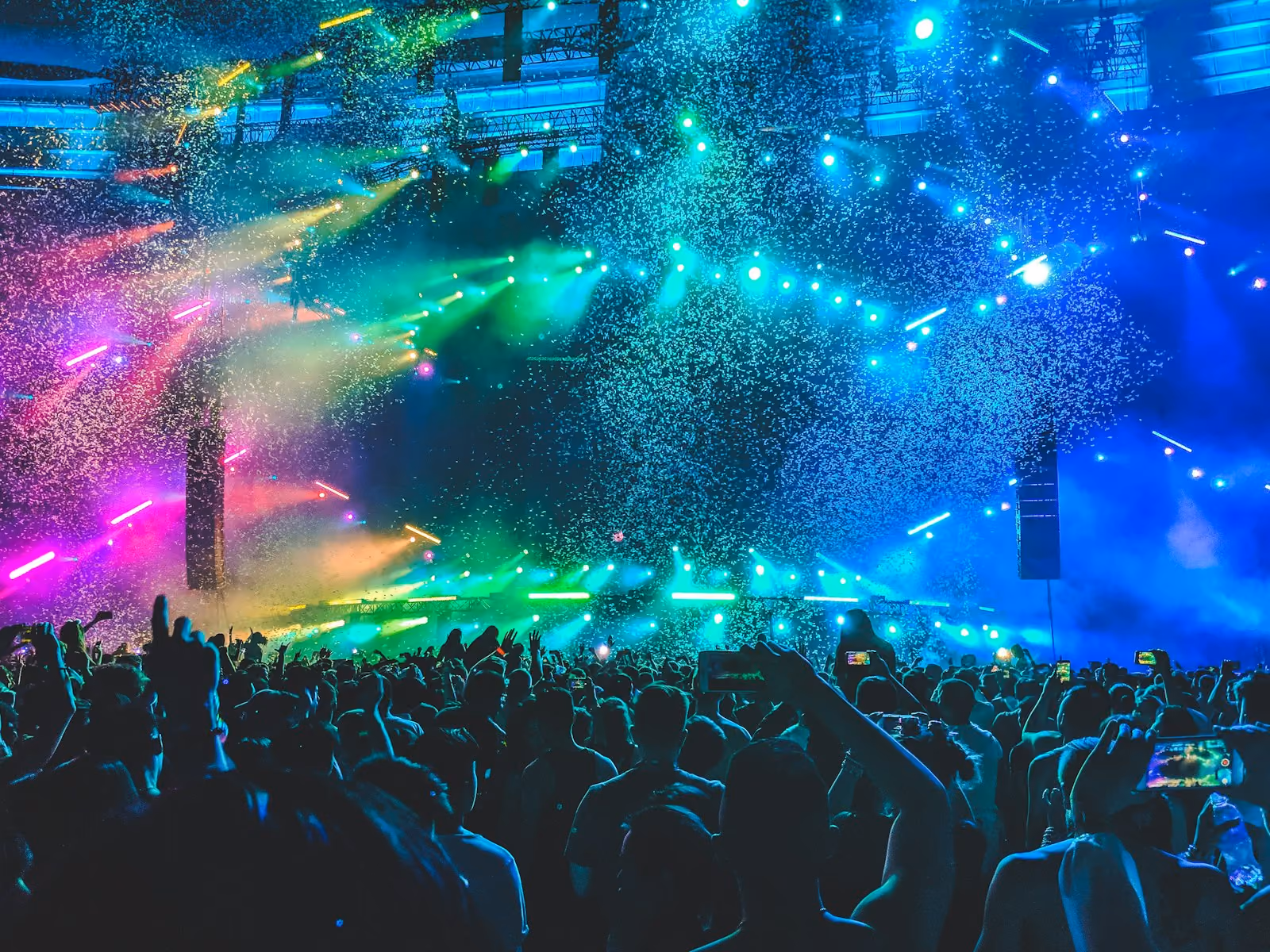 Crowd at a concert or music festival with vibrant multi-colored stage lights and confetti falling from above.