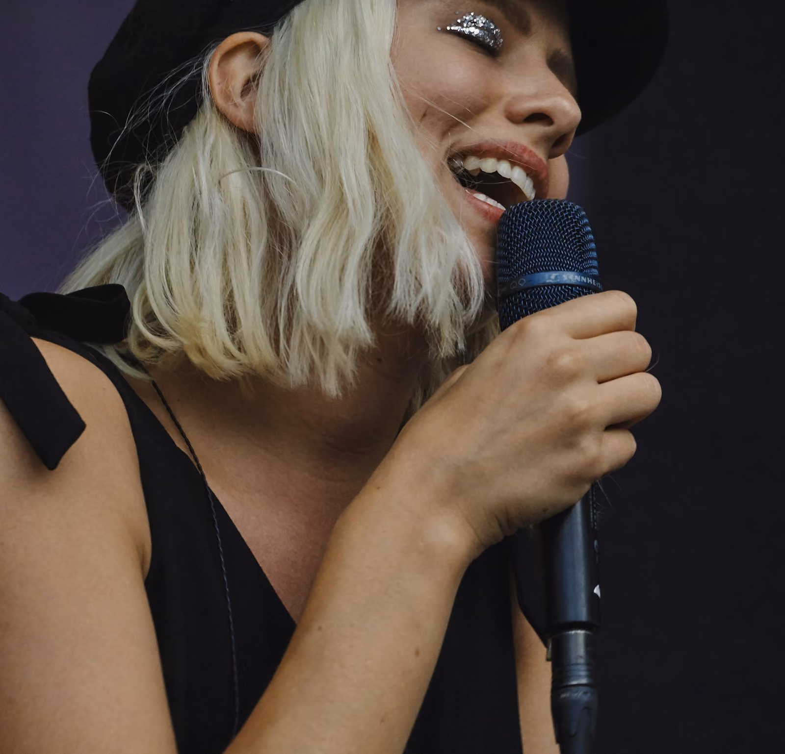 A woman with blonde hair wearing a black hat and black sleeveless top is singing passionately into a microphone with her eyes closed and glittery silver eye makeup on.