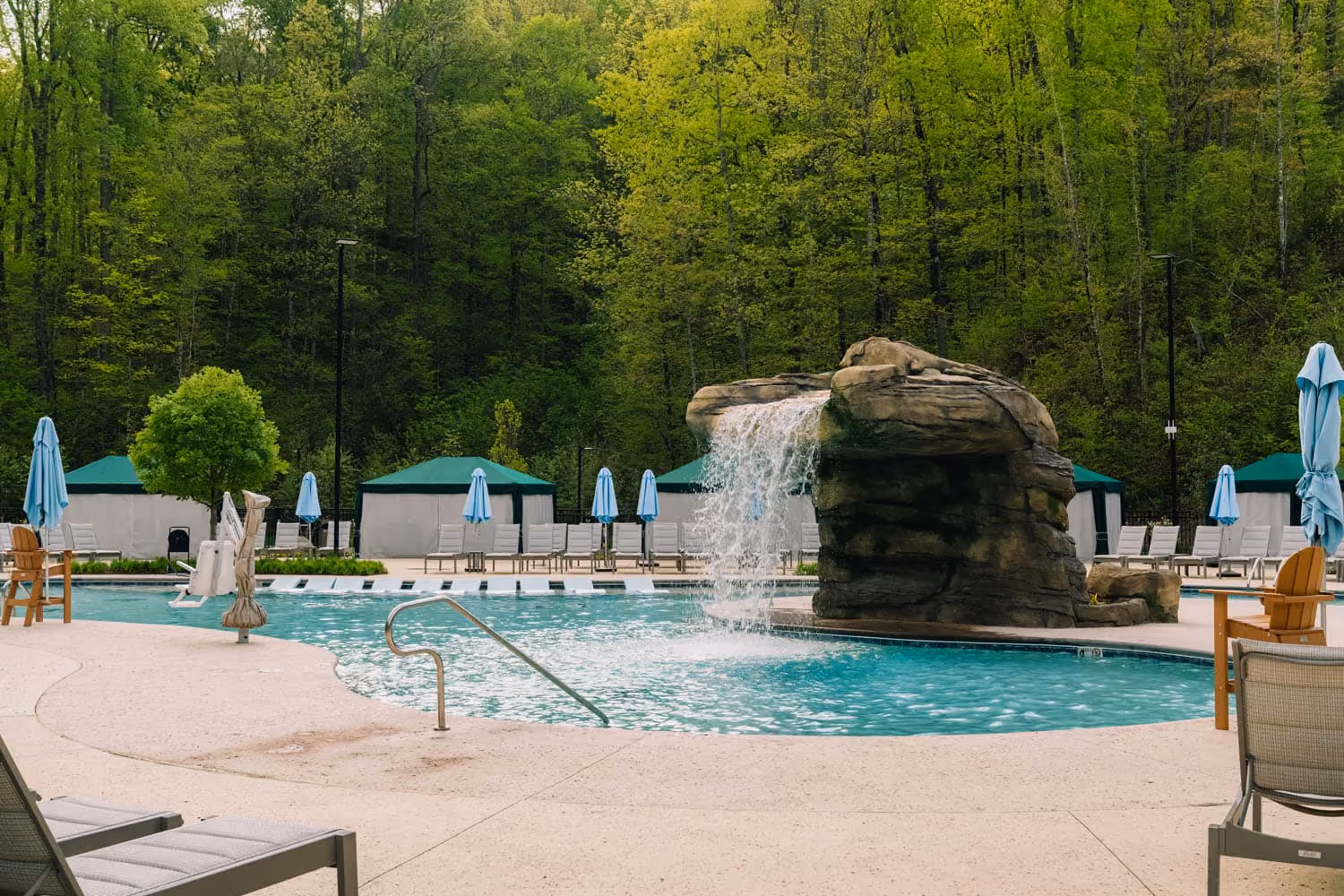 Outdoor resort pool with a rock waterfall and lounge chairs amid wooded surroundings
