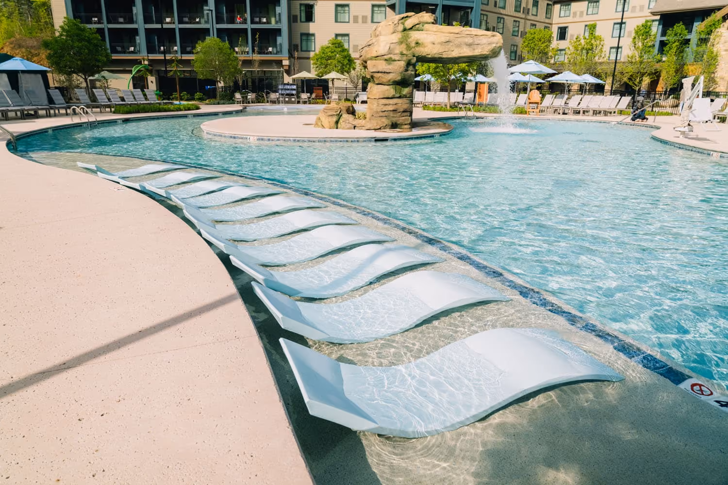 Resort-style pool with a rock waterfall and curved in-water lounge chairs at a hospitality construction site.