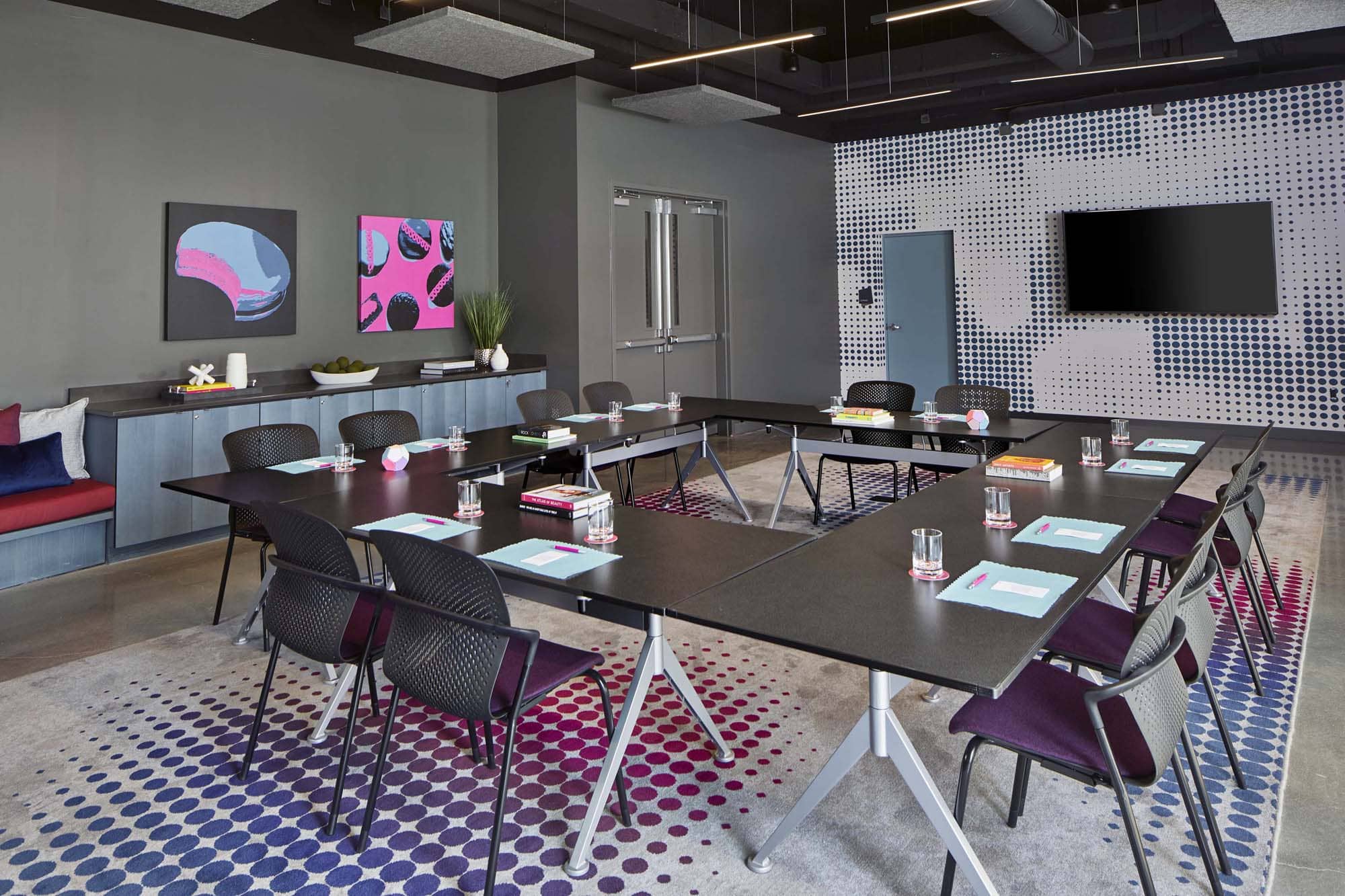 Modern hospitality meeting room with U-shaped tables, chairs, wall art, and a mounted TV