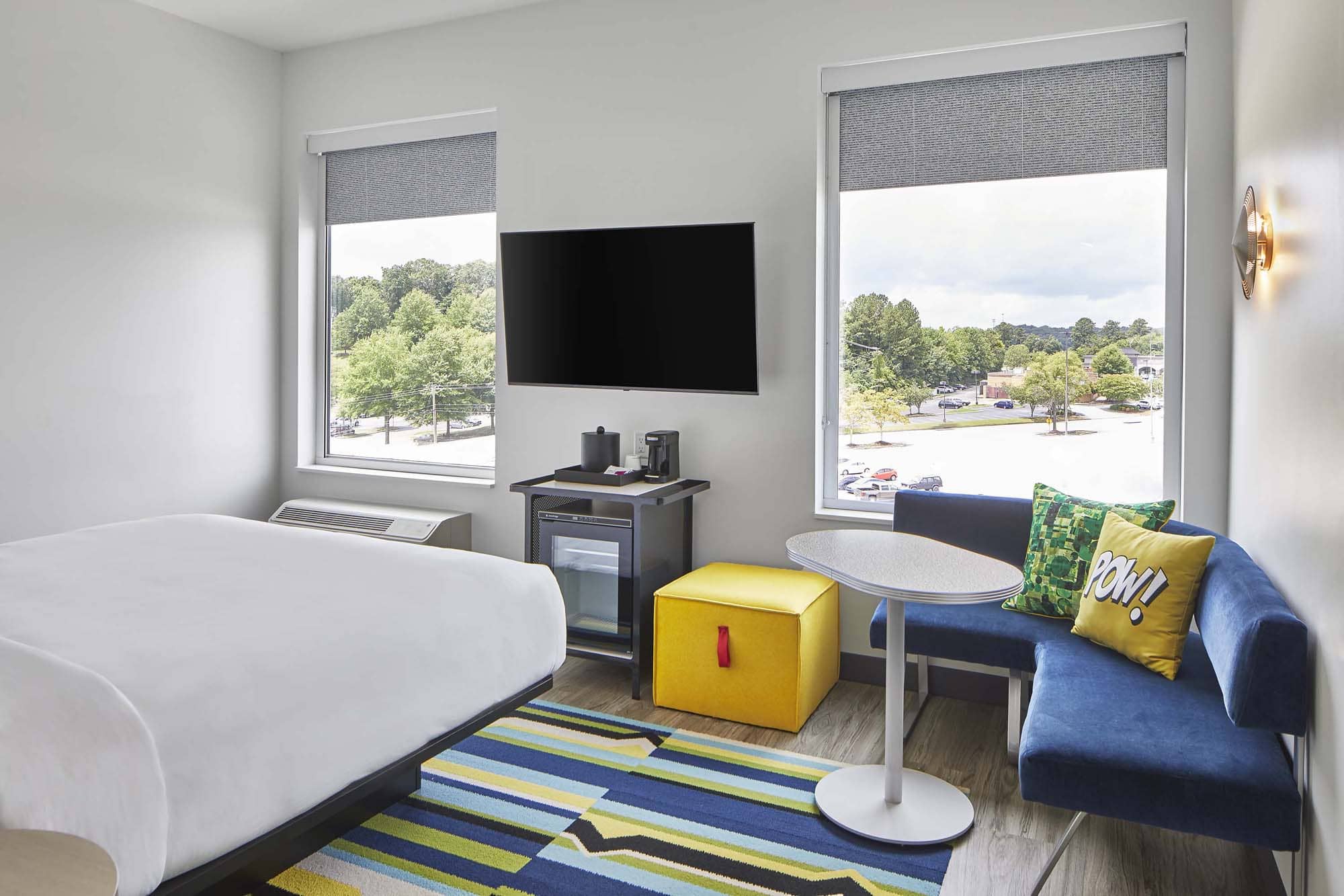 Modern hotel room with bed, TV, blue seating nook, and large windows overlooking trees and parking lot