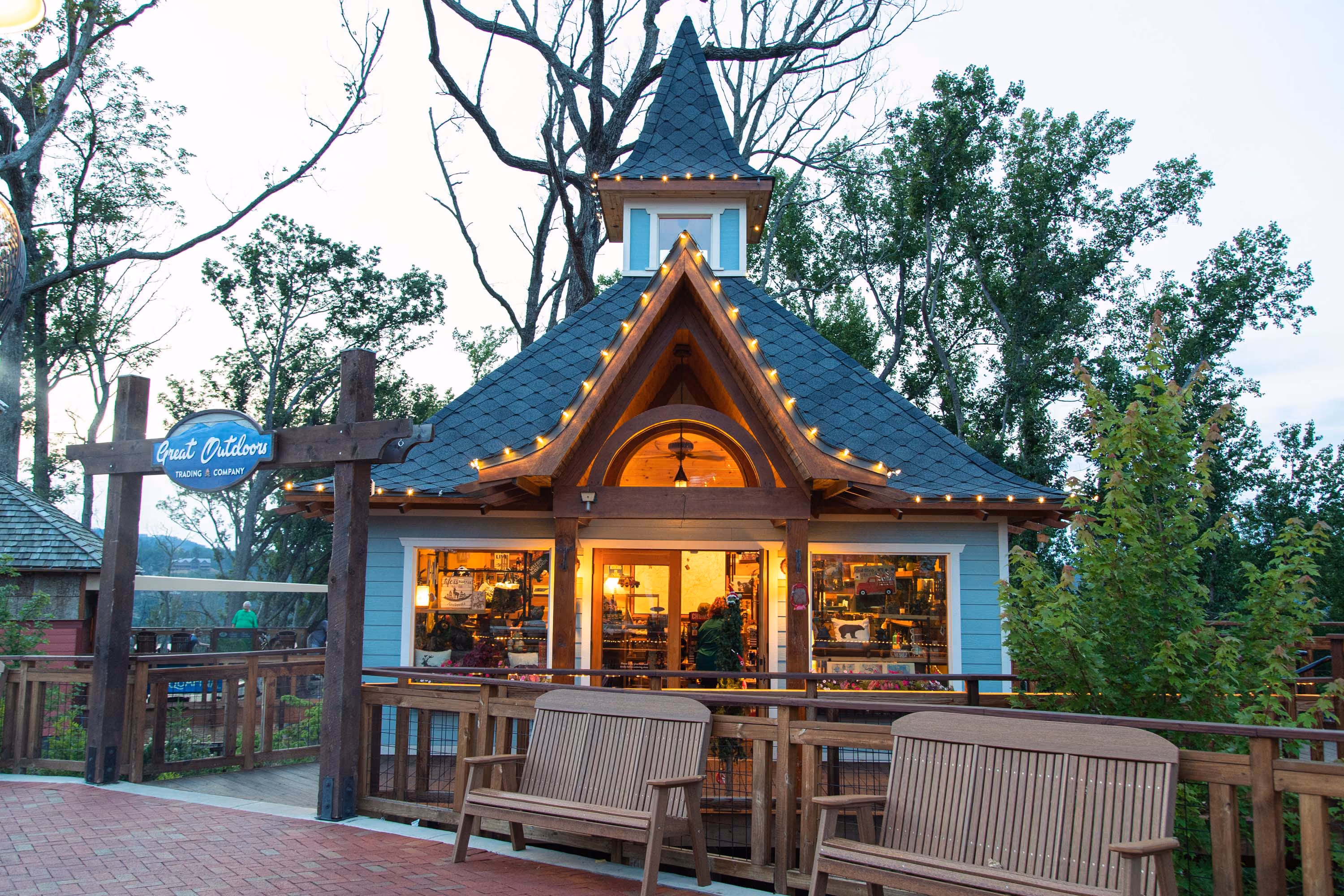 Lit-up shop with a steep roof and tower, surrounded by trees and a wooden deck at dusk.