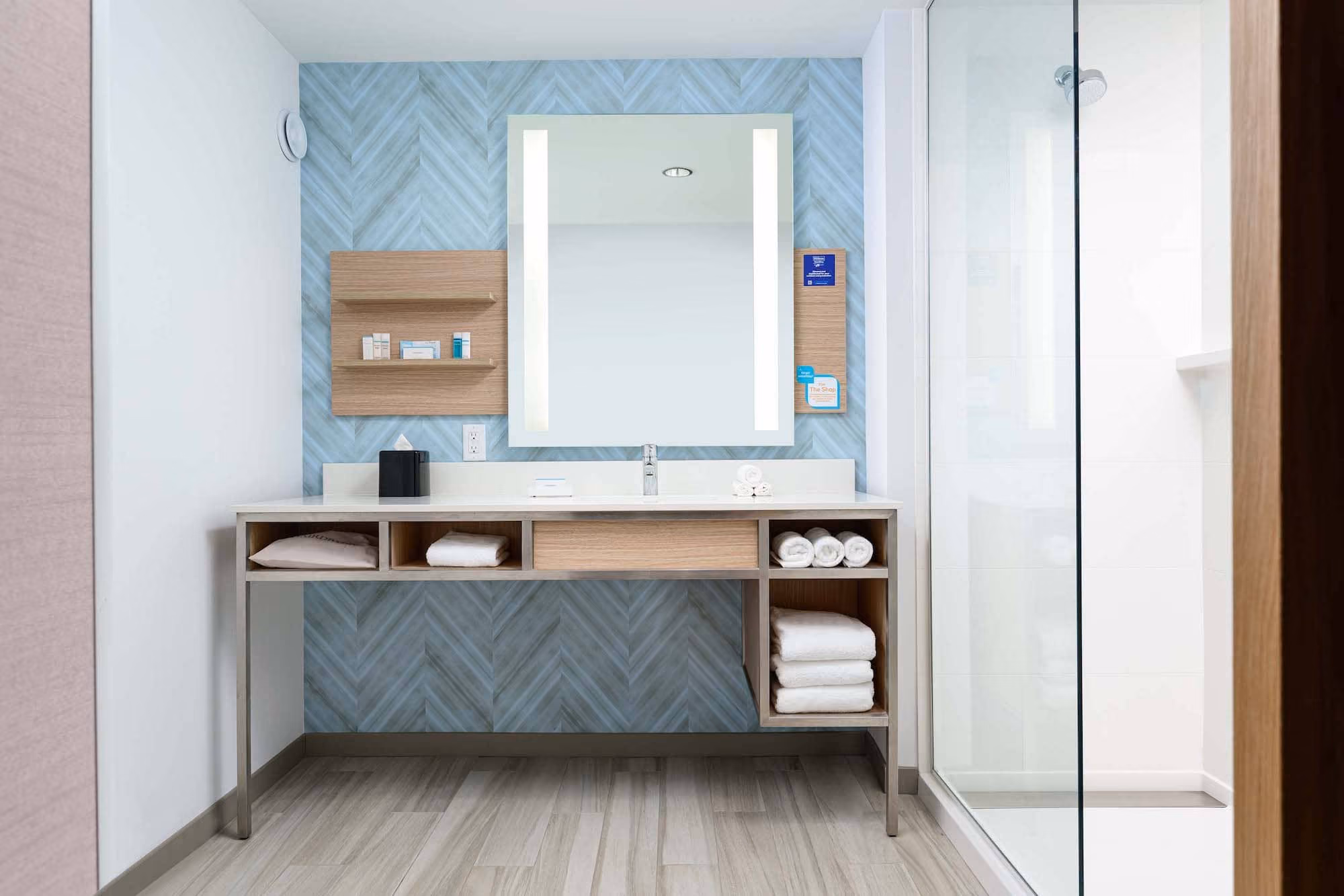 Modern hotel bathroom vanity with mirror, open shelving, and glass shower enclosure