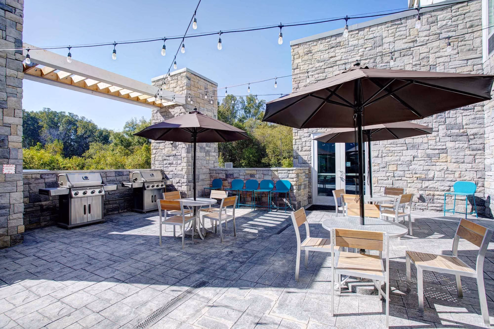 Stone patio with grills, tables, umbrellas, and string lights at a hospitality space by D&S Builders