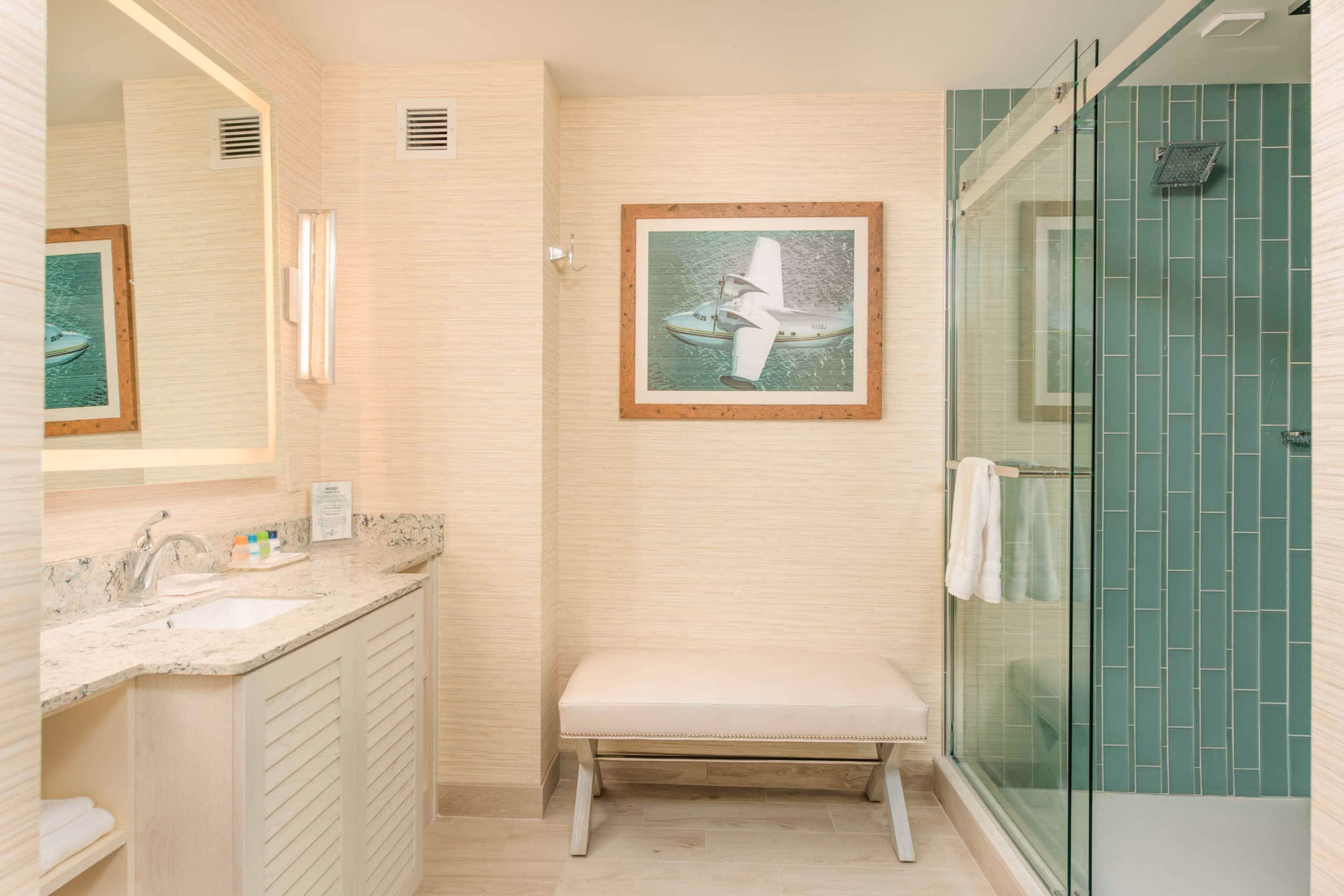 Bright hotel bathroom with vanity, bench, and glass shower with teal tile, by D&S Builders