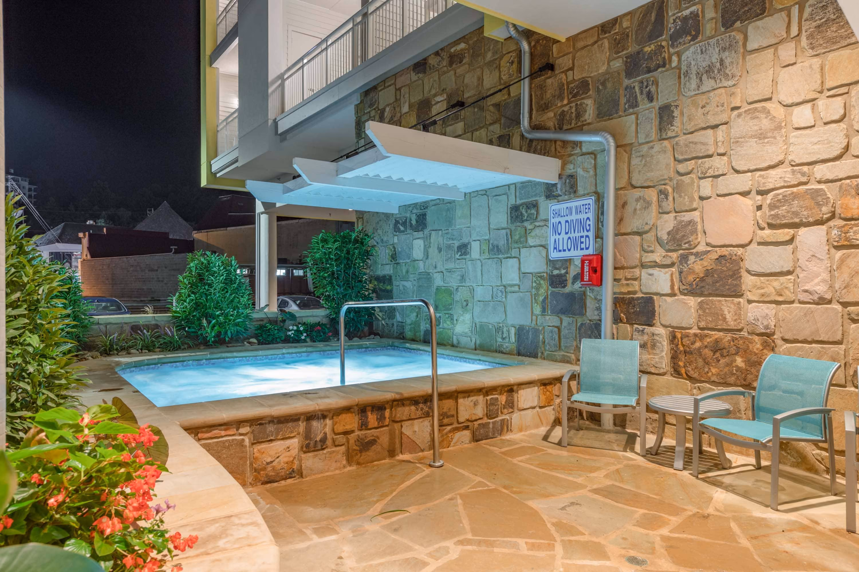 Outdoor hot tub beside a stone wall with no-diving sign and patio chairs at night